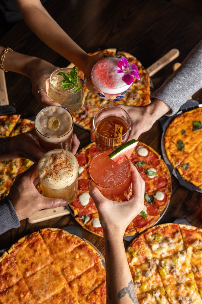 Several tavern-cut pizzas seen from above while six people reach across them
                    with cocktails to toast.