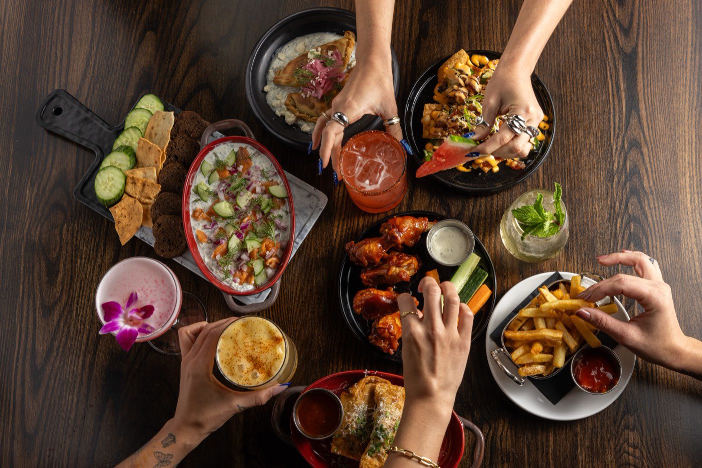 An overhead view of a full table: salmon dip with cucumber and rye crisps, gyro
                  empanadas, tuna nachos, BBQ chicken wings with crudite and ranch, fries, and
                  cocktails, with several people reaching in at once.