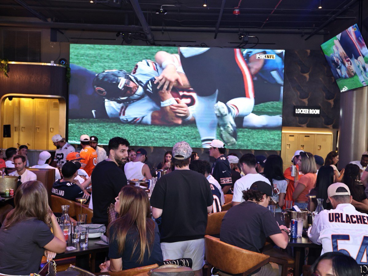 The seventeen-foot LED screen carrying a Bears game over a packed room.