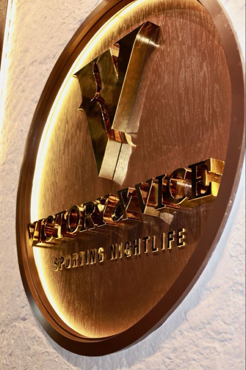 The brass Victory & Vice sporting nightlife medallion lit against white brick.