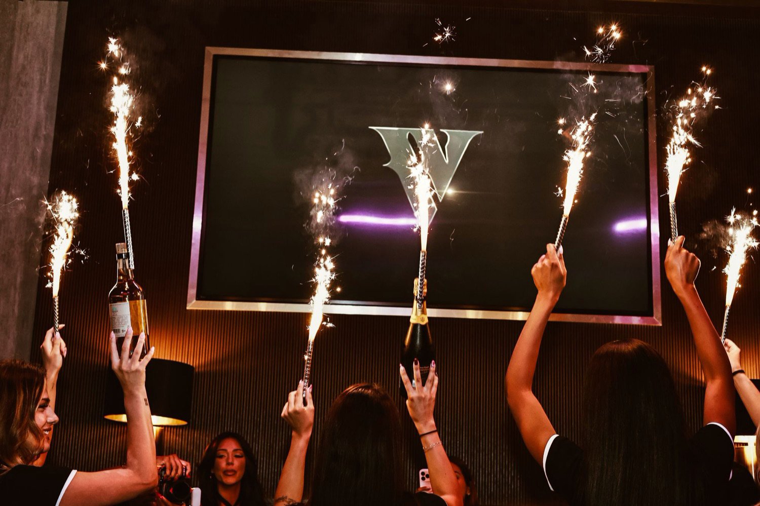 Sparklers raised together beneath the lit V screen as bottles arrive at the table.