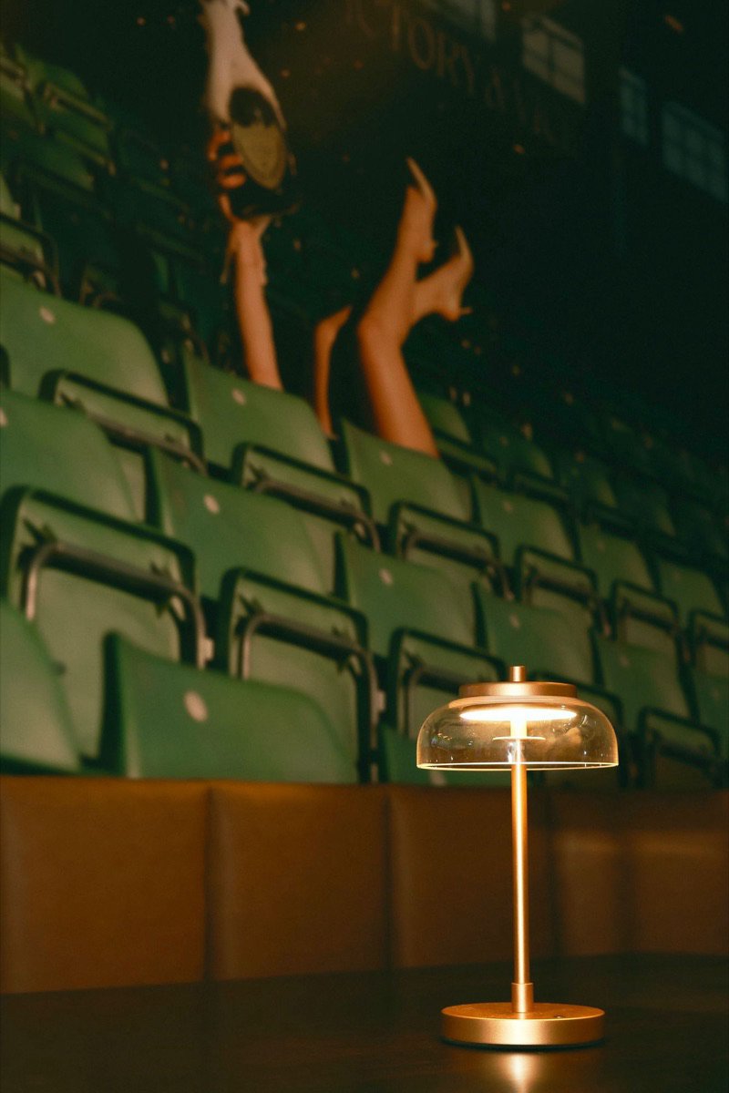 A brass table lamp lit on the rail, stadium seating on the wall behind it.