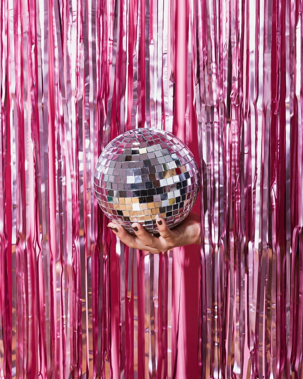 A hand holding a mirrored disco ball against a curtain of pink tinsel.