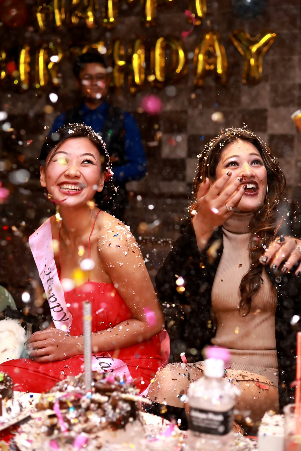 Two friends laughing under falling glitter at a birthday party, one wearing a birthday sash.